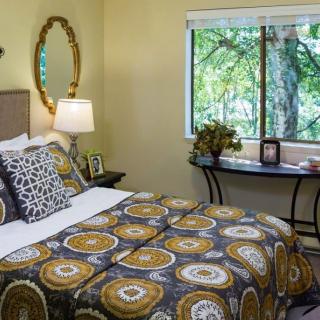 A cozy bedroom features patterned bedding, a decorative mirror, and a window with a view of greenery.