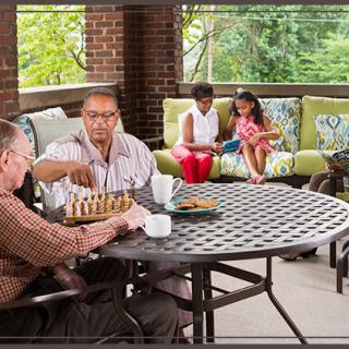 Family relaxing on a porch, with two men playing chess and a mother and daughter sitting on a couch.