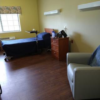 An interior view of a hospital or rehabilitation room featuring a bed, chair, and storage.