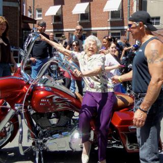 An elderly woman joyfully poses on a motorcycle during a community event, surrounded by onlookers.