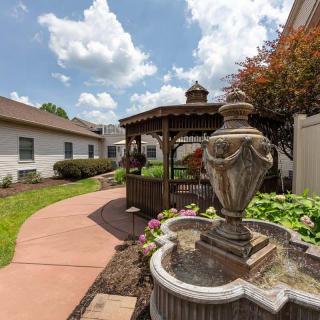 A scenic garden with a decorative fountain and wooden gazebo under a partly cloudy sky.