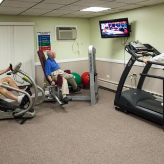 A small indoor gym with people using exercise equipment while watching television.