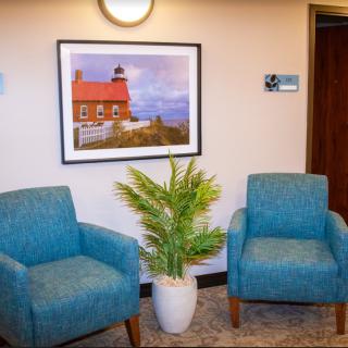A cozy waiting area featuring two blue chairs, a potted plant, and a framed lighthouse photograph on the wall.