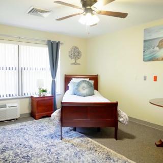 A cozy bedroom with a single bed, nightstand, and ceiling fan, featuring modern decor and natural light from a large window.