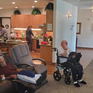A cozy nursing home setting with elderly residents relaxing and caregivers working in the kitchen.