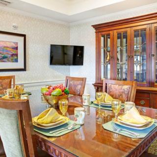 A well-appointed dining room with a polished wooden table, elegantly set with place settings and a fruit bowl, complemented by a china cabinet and wall-mounted television.