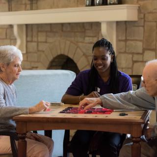 A caregiver spending time with two elderly individuals playing a board game.
