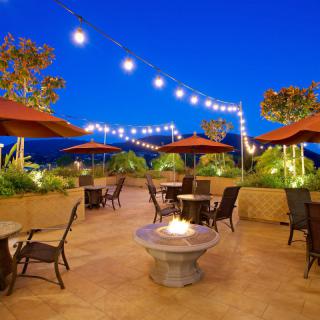 A cozy outdoor patio with string lights and a fire pit, featuring tables and chairs under umbrellas during the evening.