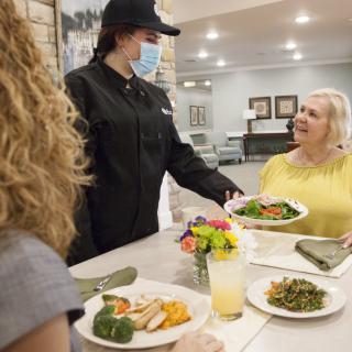 A waitress wearing a face mask serves food to seated customers in a dining setting.