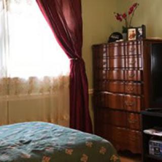 A cozy bedroom featuring a window with red curtains, wooden dresser, television, and decorated bed.