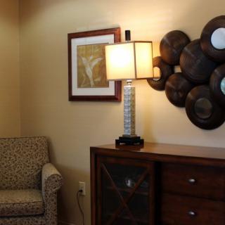 A cozy living room corner featuring a patterned armchair, a lamp on a wooden cabinet, and wall art.
