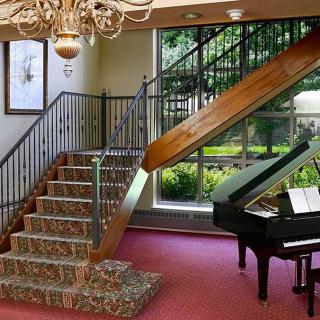 A luxurious interior featuring a grand staircase with a patterned carpet, a grand piano by the window, and an elegant chandelier overhead.