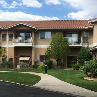 A two-story residential apartment building with a landscaped yard, featuring trees and a grassy area under a partly cloudy sky.