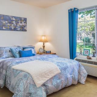 A cozy bedroom with blue accents, featuring a bed, nightstand with lamp, and a window with blue curtains.