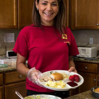 A woman in a red shirt smiling while serving a meal in the kitchen.