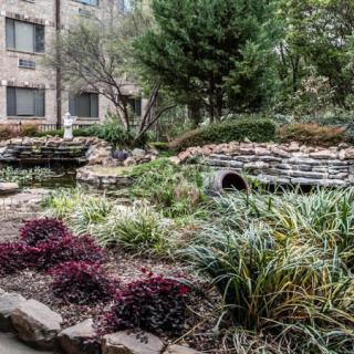 A serene garden with lush greenery, stone pathways, and a tranquil pond amidst an urban setting.