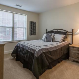 A cozy bedroom featuring a neatly made bed with matching linens, a bedside lamp on a wooden nightstand, and a window allowing natural light to illuminate the room.