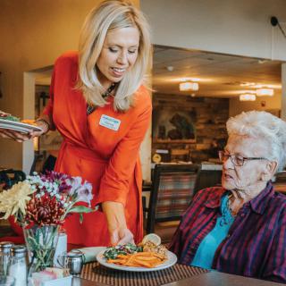 A caregiver serves a meal to an elderly person at a dining table.