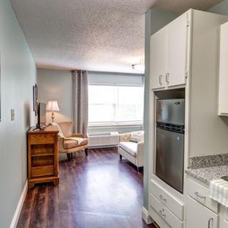A cozy apartment interior with a small kitchen leading to a bright living room featuring comfortable furniture and a large window.