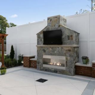 A modern outdoor patio featuring a stone fireplace with a mounted TV and surrounding landscaping.