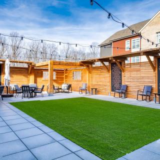A modern backyard with a patio featuring artificial grass, string lights, and seating under a pergola.