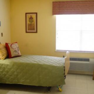 A cozy bedroom with a single bed featuring a green bedspread and decorative pillows, complemented by a window with blinds and wall art.