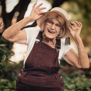An elderly woman in overalls joyfully poses with her hat while enjoying the outdoors.