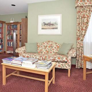 A cozy living room with a floral patterned sofa, a bookshelf filled with books, and a coffee table topped with magazines. The room features green walls, a decorative painting, and matching curtains.