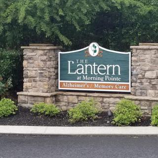 A sign for The Lantern at Morning Pointe, a memory care facility, mounted on a brick wall surrounded by greenery.