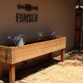 A wooden raised garden bed on a patio, accompanied by watering cans and a 'Family' sign on the wall.