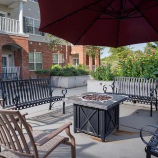 A cozy outdoor patio with a fire pit, surrounded by benches and chairs under a large umbrella, adjacent to a residential building.