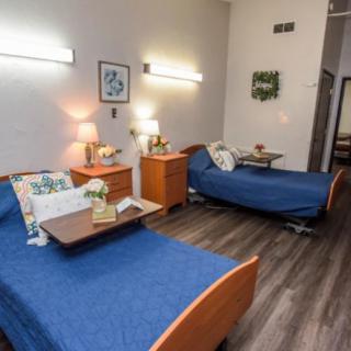 A hospital room featuring two neatly made beds with blue bedding, accentuated by lamps and side tables on a wooden floor.