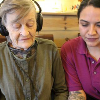 An elderly woman wearing headphones sits next to a young woman indoors.