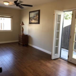 A spacious living room with hardwood floors, featuring a ceiling fan, French doors leading outside, a window, and decorated with a painting and a sculpture.