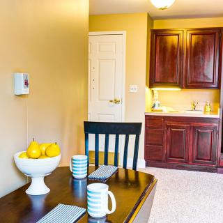 A cozy dining area next to a kitchen, featuring a dark wooden table with striped mugs and a fruit bowl filled with lemons.