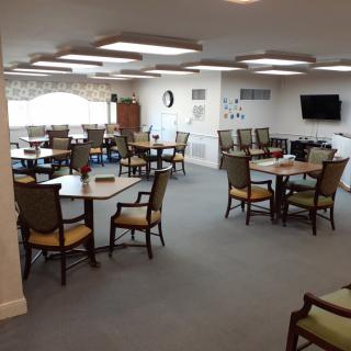 An interior view of a dining area with multiple tables and chairs, a wall-mounted television, and large windows.