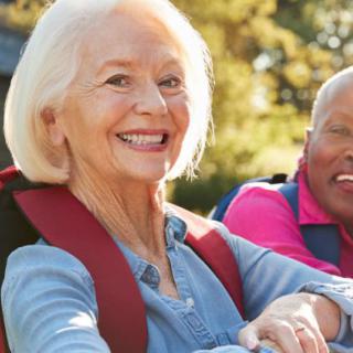 An elderly couple enjoying a hike outdoors, both smiling and wearing backpacks.