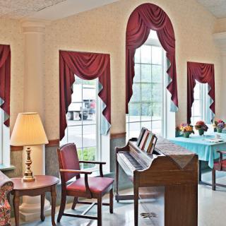 A cozy interior with a piano, chairs, and large windows adorned with red draped curtains.