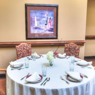 A round dining table set for four with white tablecloth and elegant place settings, positioned beneath a framed painting in a warmly lit room.