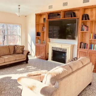 A cozy living room with two sofas facing a fireplace and a large television, surrounded by built-in bookshelves and sunlight streaming through the windows.