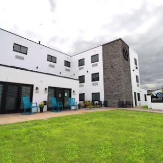 A modern building with a mix of white and stone exterior, featuring outdoor seating on a grassy area under a cloudy sky.