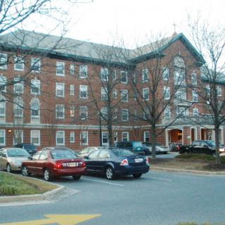A four-story brick residential building with a parking lot in front, surrounded by trees.