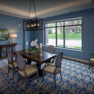 A stylish dining room with blue walls, a wooden table, and upholstered chairs, featuring a modern chandelier and a scenic window view.