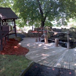 A peaceful garden setting with a wooden walkway leading to a gazebo under lush trees.