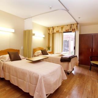 A hospital room with two beds, wooden flooring, and natural light from a window.
