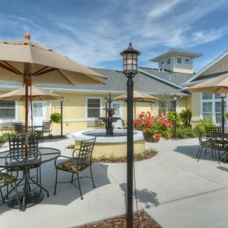 A sunny patio area with tables, chairs, and umbrellas surrounding a central fountain in front of a building.