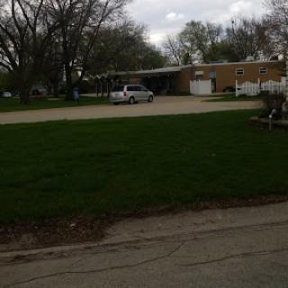 A quiet suburban scene featuring a house, green lawn, and parked car under a cloudy sky.