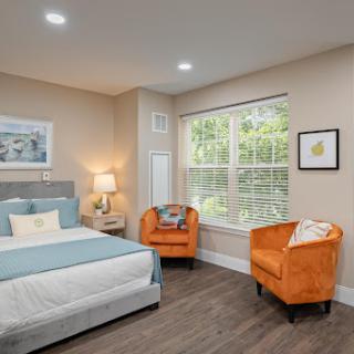 A cozy and modern bedroom featuring a comfortable bed, vibrant orange chairs, and ample natural light from the large window.