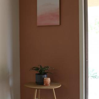 A minimalist interior featuring a small wooden table with a plant and candle, set against a brown wall with abstract art.