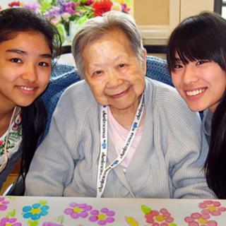 An elderly woman smiles warmly while seated between two younger women, representing multiple generations and family togetherness.